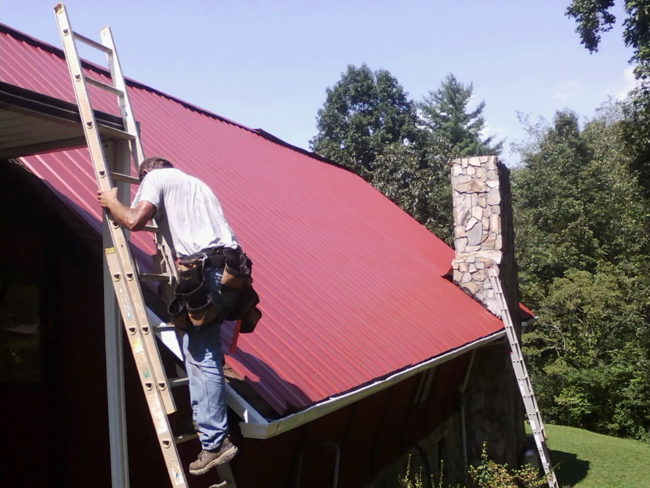 Roofer on a ladder beside a red metal roof and stone chimney — Soffit & Fascia Repair by Roofing Brothers in Lincolndale